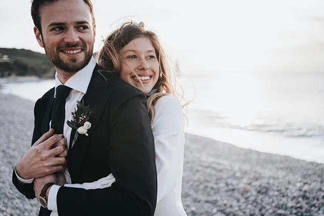 Un couple heureux dans des costumes de mariage classiques contre un décor de plage ensoleillé, idéal pour un mariage en