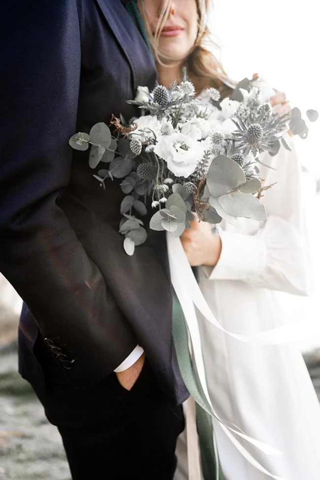 Un couple de mariée et de mari en tenue élégante tenant un bouquet romantique de fleurs grises et blanches.