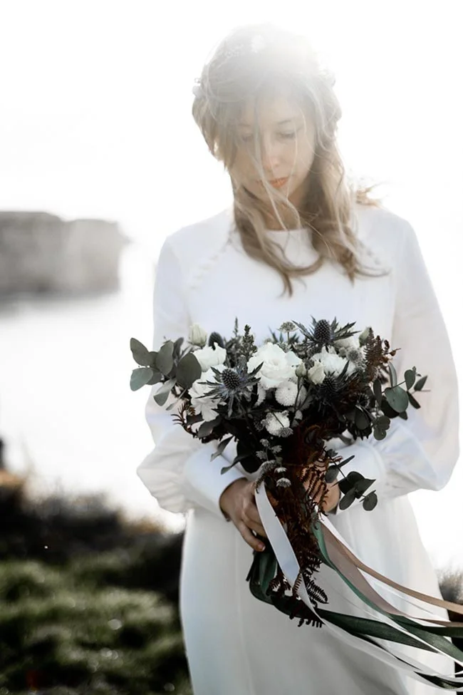 Une mariée en robe blanche tendre tenant un bouquet romantique de fleurs noires et blanches, avec un fond de mer douce