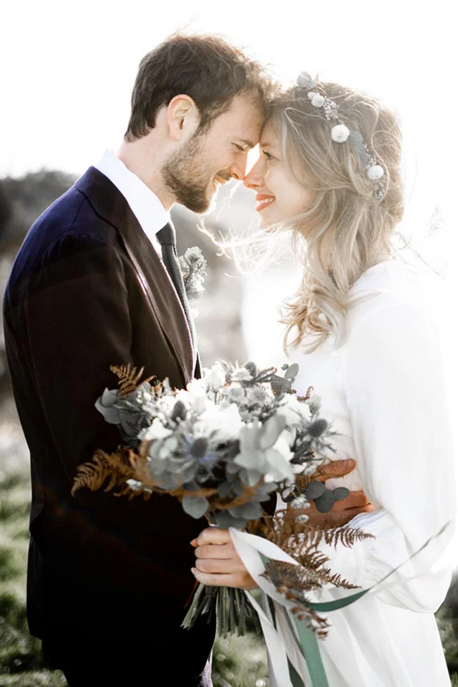 Un couple charmant se penche l'un envers l'autre, tenant un bouquet romantique de fleurs grises et dorées, sous un ciel
