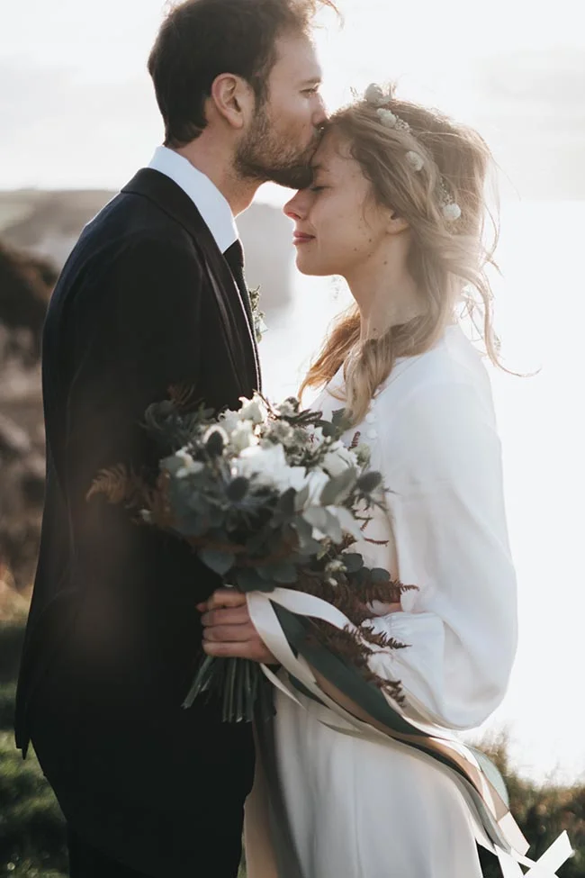 Un couple romantique partage un moment tendre au bord de la mer, le mari en costume noir, la mariée en robe blanche