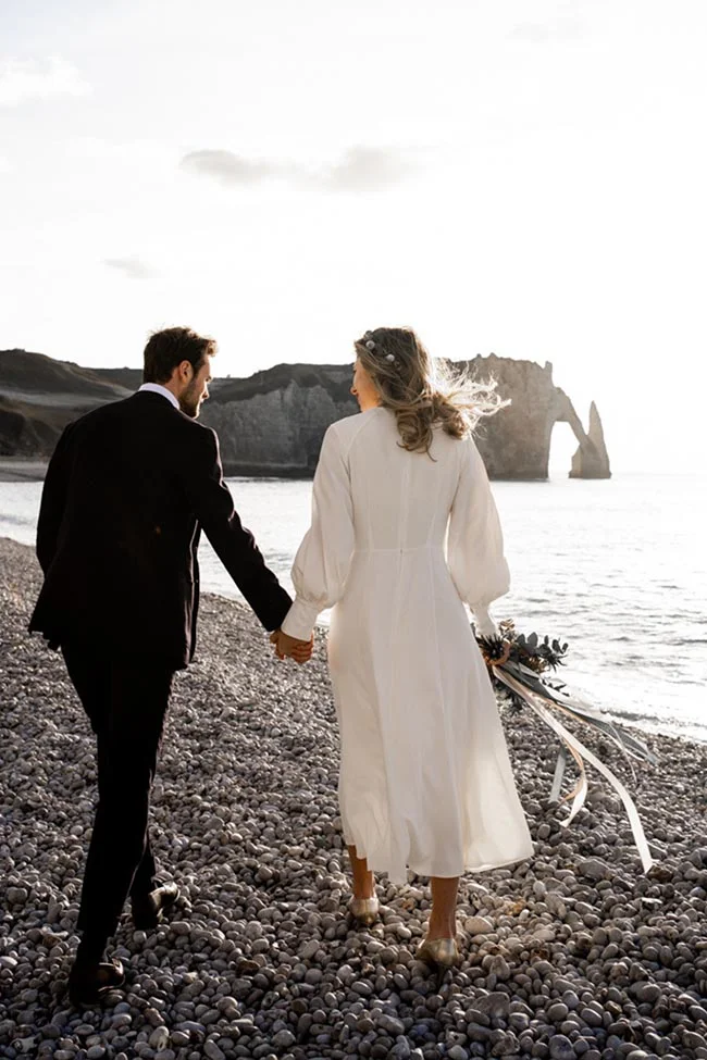 Un couple romantique se promène sur une plage rocheuse, avec les falaises d'Étretat en arrière-plan, le jour ensoleillé.