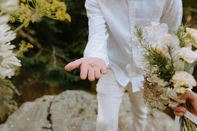 Un couple en blanc partageant des anneaux de mariage, entouré de fleurs blanches et verdoyantes dans les Pyrénées.