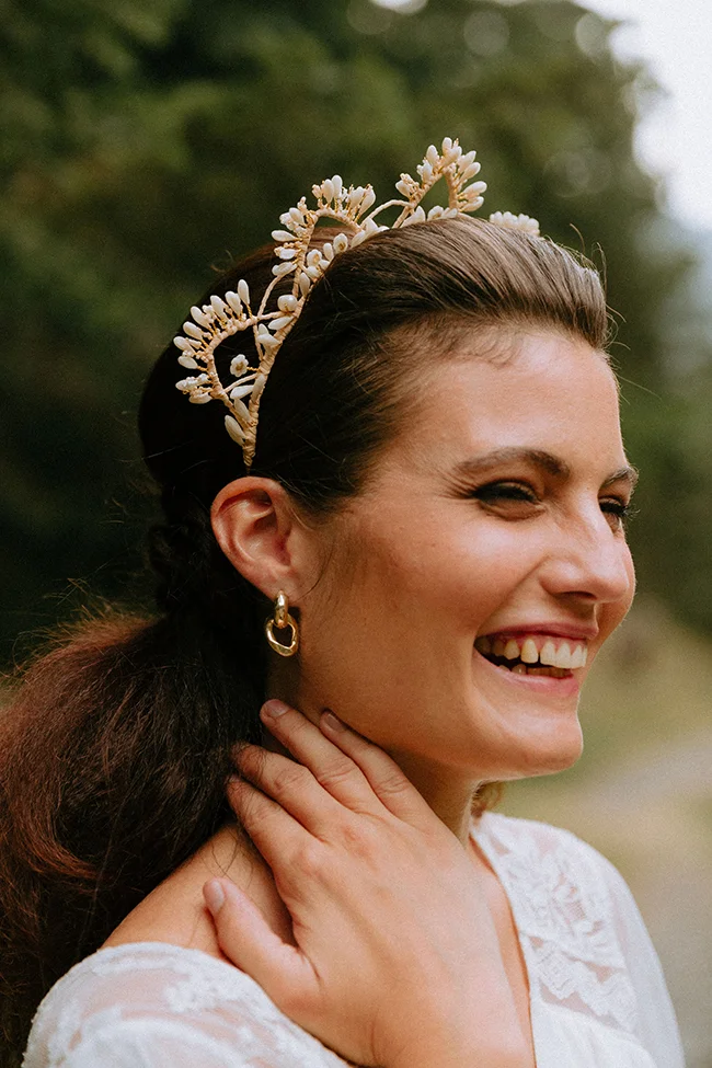 Une mariée heureuse dans les Pyrénées, ornée d'un diadème en perles blanches et d'oreilles dorées, sourit en se caressant le cou.