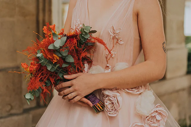 Une jeune femme dans un robe de mariage rose tient un bouquet vif de fleurs rouges et vertes, symbolisant l'amour et la joie.