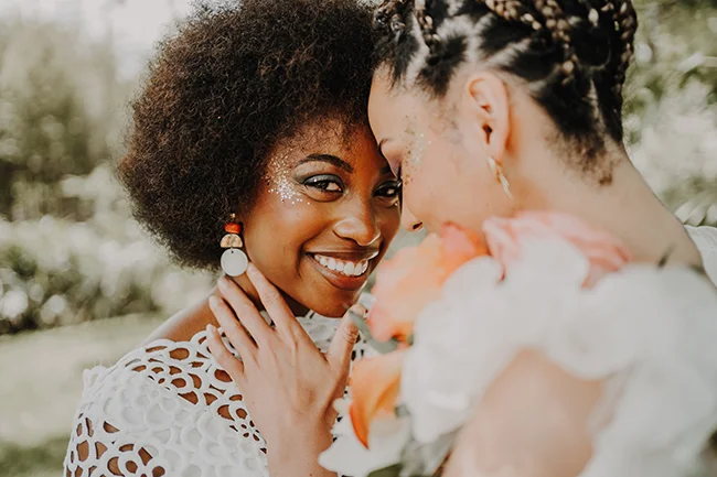 Une mariée éclatante dans un robe blanc orné de motifs floraux, se penche pour un baiser tendre avec un partenaire, au