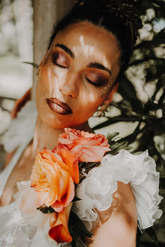 Une femme paisible au visage doré, fermant les yeux, tenant un bouquet de roses chaudes, dans un décor luxuriant.