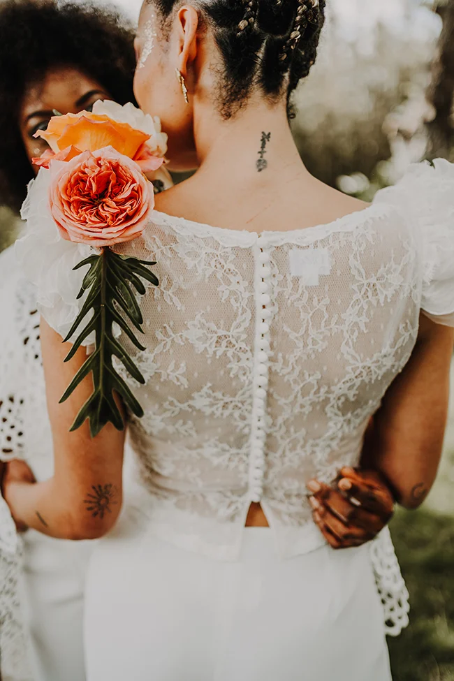 Une mariée en robe blanche ornée de fleurs roses et de motifs floraux, avec tatouages étonnants, dans un décor exotique.