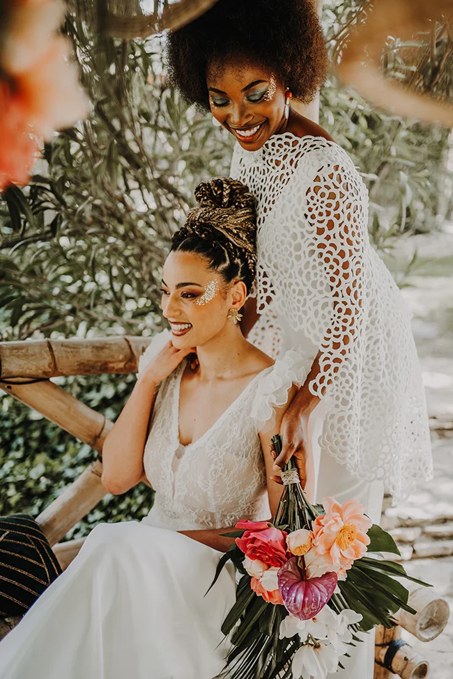 Une mariée en blanc et une amie aux cheveux afro dans un décor exotique, avec une bouquet coloré de fleurs.