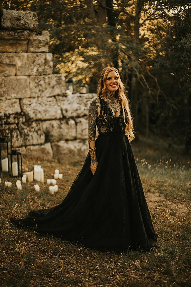 Une jeune femme dans un long robe noir éclatante de tatouages se tient dans un décor romantique avec des bougies,
