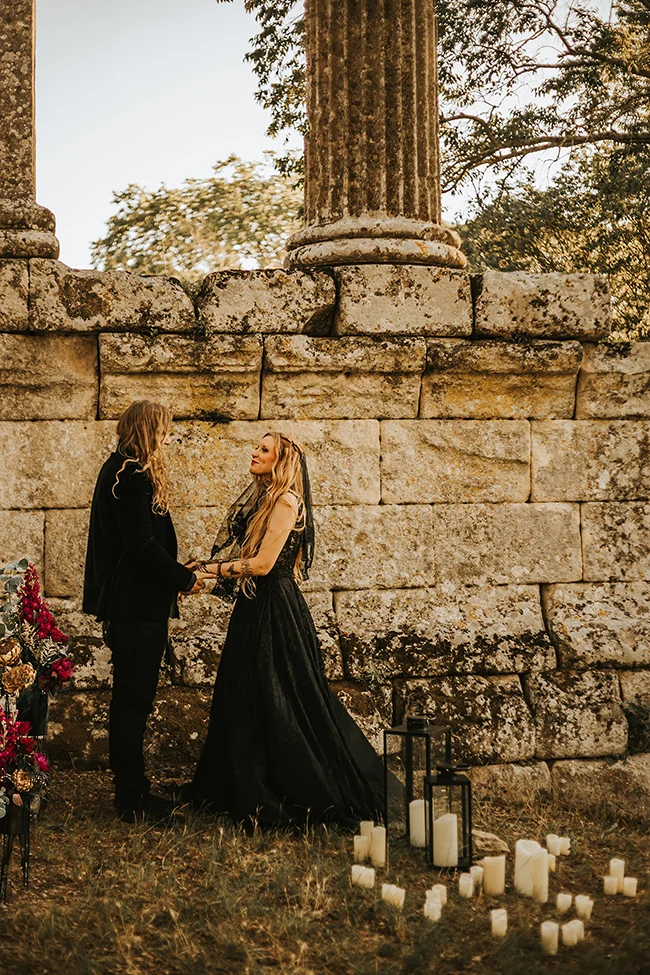 Un couple élopé échange des vœux dans un décor romantique et mystérieux, avec des bougies et des fleurs noires en témoignage.