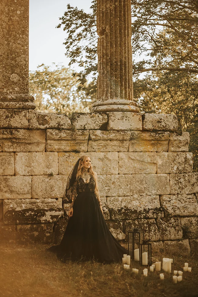 Une jeune femme dans un vêtement sombre et élégant se tient devant des colonnes anciennes, entourée de candelles,