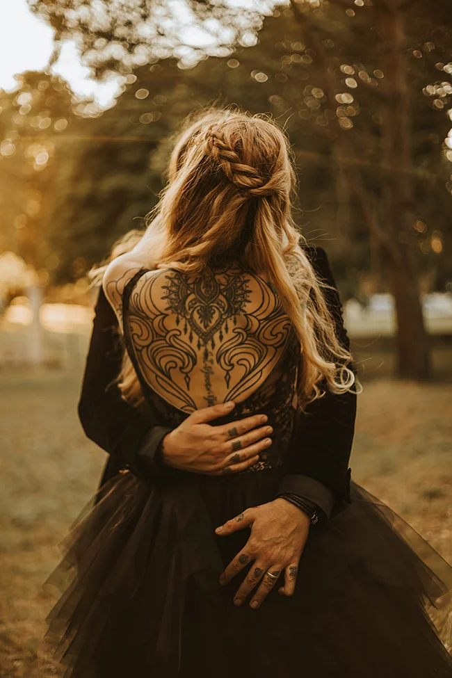 Une mariée éloignée dans un décor romantique, son dos orné d'un tattoo mystérieux, habillée d'un robe sombre et tourbillonnante.