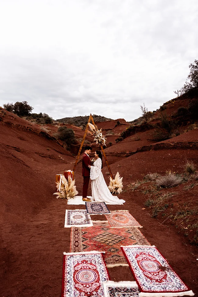 Un couple élopé se tient sous un arc décoratif dans un canyon coloré, entouré de tapis traditionnels et de décorations bohèmes.
