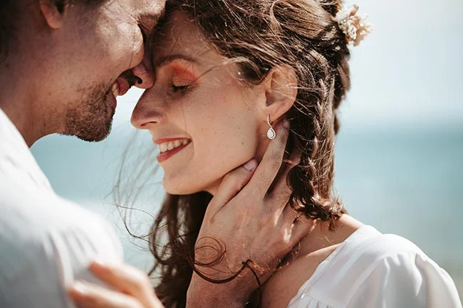Une jeune mariée dans un vêtement blanc tendre se penche pour un baiser tendre avec son mari, contre un fond de mer serein.
