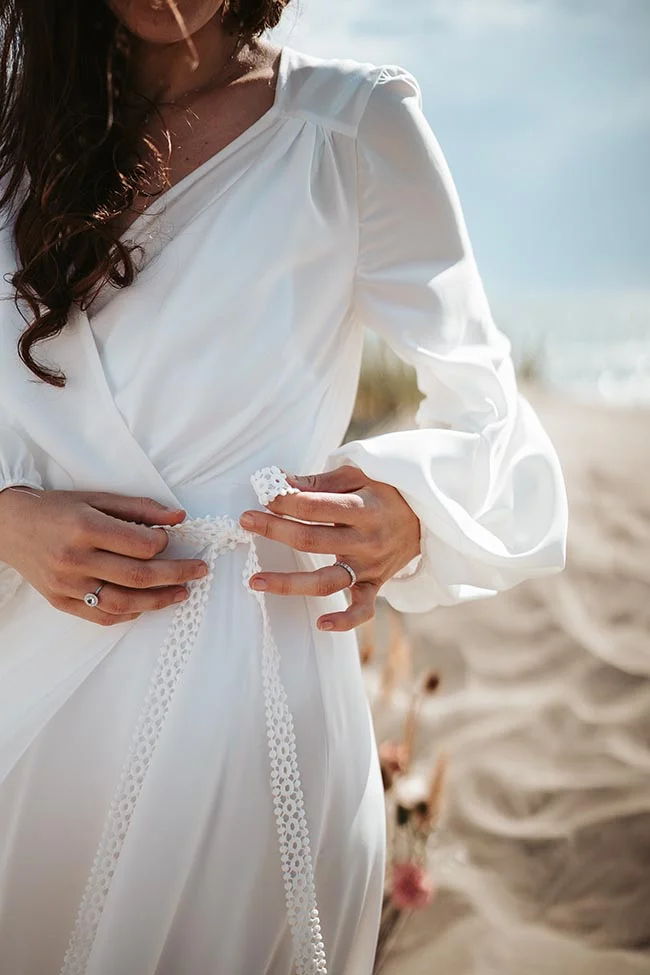 Magnifique bride en robe blanche légère s'empare de son corset, bordé de dentelle, sur une plage paisible.