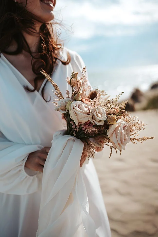 Une mariée blanche tenant un bouquet romantique de fleurs blanches et brunes sur une plage paisible.