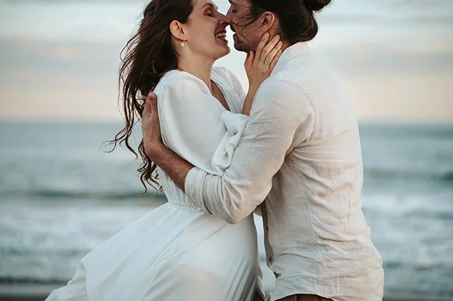 Un couple heureux s'embrassant tendrement sur la plage, vêtu de blanc, capturant l'ambiance romantique d'un elopement