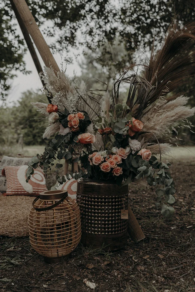 Un bouquet de fleurs bohémienne avec des roses et des plumes, accompagné d'un lampadaire en rattan, évoquant un mariage