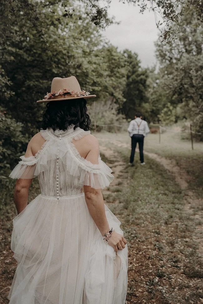 Une mariée en robe blanche bohème et chapeau fleuri s'approche d'un marié en costume vintage dans un parc charmant.