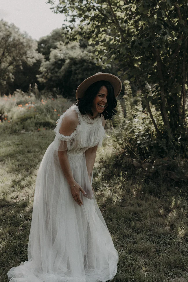 Une mariée en robe blanche légère et élégante se penche dans un champ verdoyant, souriant, pour un instantané