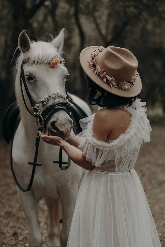 Une jeune femme en robe blanche tend la main pour caresser un cheval blanc orné de fleurs, dans un décor paisible et romantique.