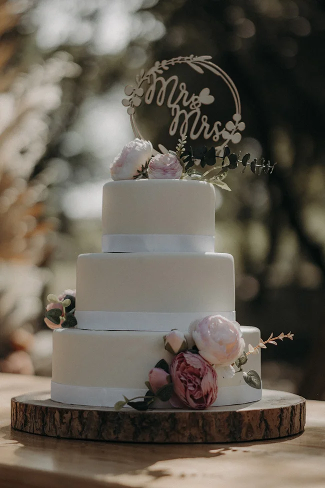 Un gâteau de mariage bohémien avec une toche "Mr & Mrs" élégante, décoré de fleurs roses et blanches, reposant sur un