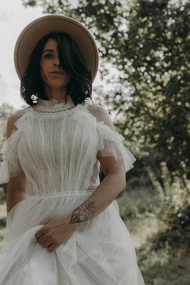 Une jeune femme dans un robe blanche bohémienne et un chapeau large, avec une tattoo étonnante sur le bras, se tient