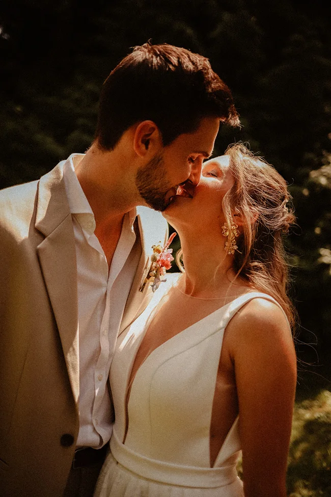 Un couple de mariée et de mari en blanc et beige partageant un moment tendre dans un décor verdoyant.