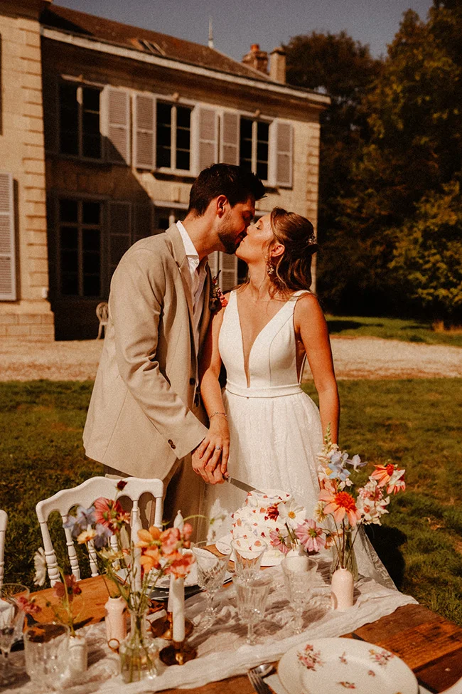 Un couple romantique se coupe un baiser tendre au fond d'un décor intime et coloré, avec des fleurs et un repas soigné.