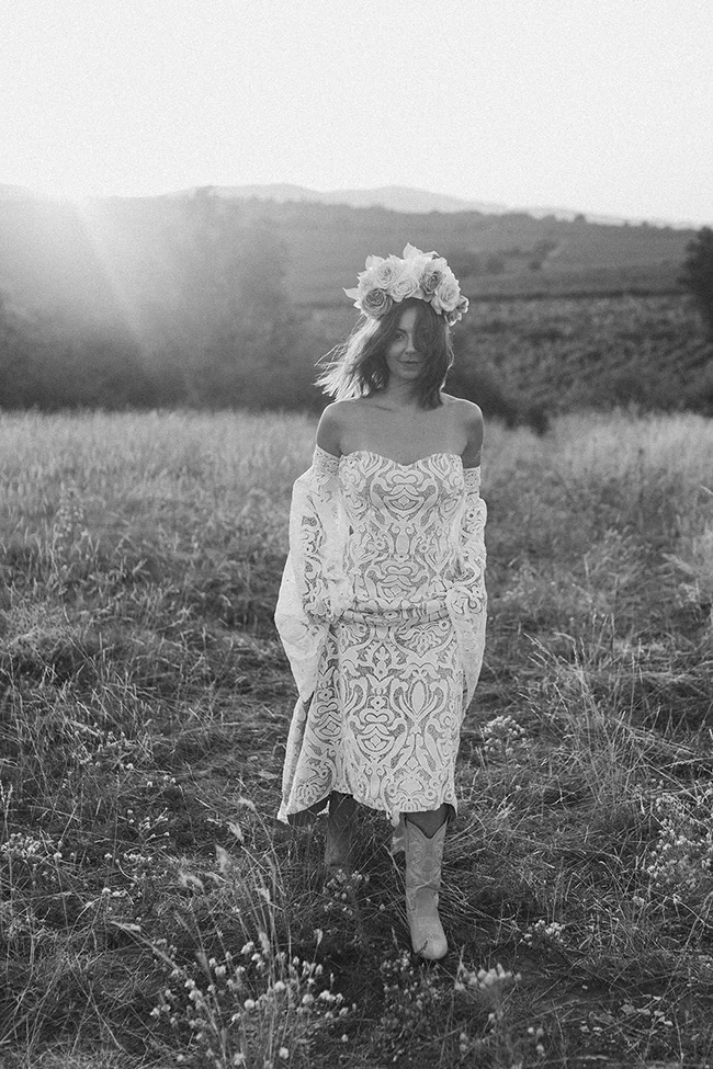 Élégante mariée en robe décolleté ornée de motifs indiens, couronnée de fleurs, dans un champ herbeux au coucher de soleil.