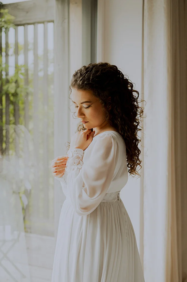 Une jeune femme dans un robe blanche élégante se tient devant une fenêtre, évoquant un sentiment de tranquillité et de réflexion.