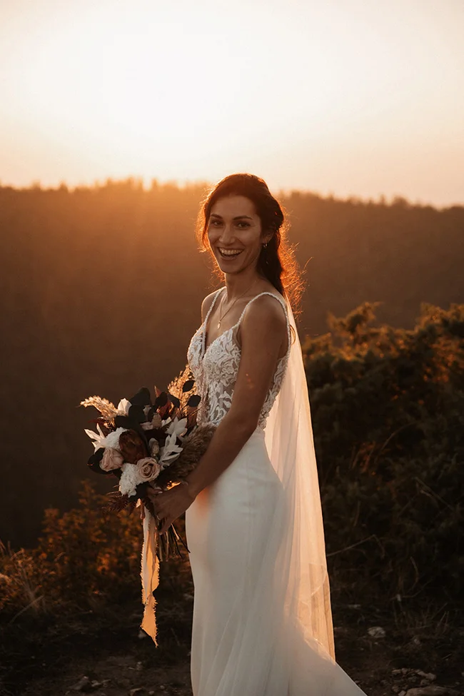 Une mariée heureuse tenant un bouquet romantique, contre un fond d'or du coucher de soleil, dans le parc national du Vercors.