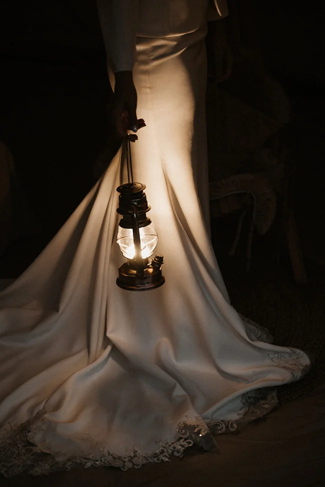 Élégante mariée tenant un lampion vintage, éclairant son robe blanche et son train orné de dentelle, dans un décor