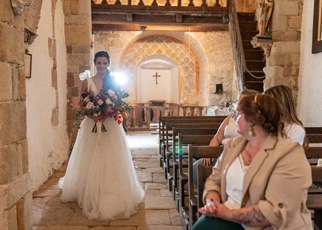 Une mariée dans une robe blanche traditionnelle tient un bouquet de fleurs dans un église de charme, avec des