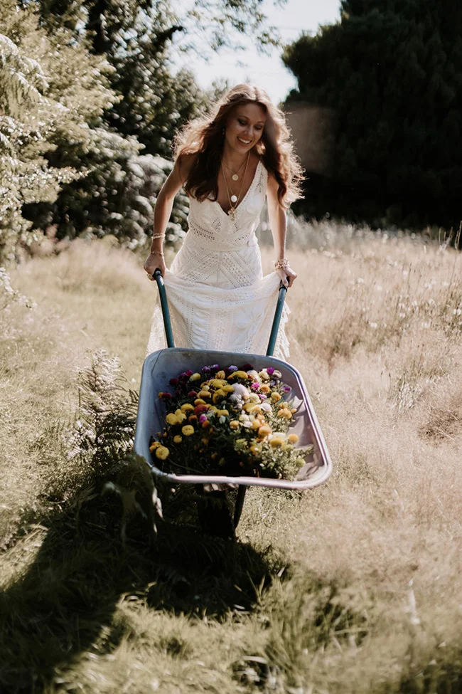 Une mariée souriante en robe blanche poussant une brouette remplie de fleurs colorées dans un champ ensoleillé.