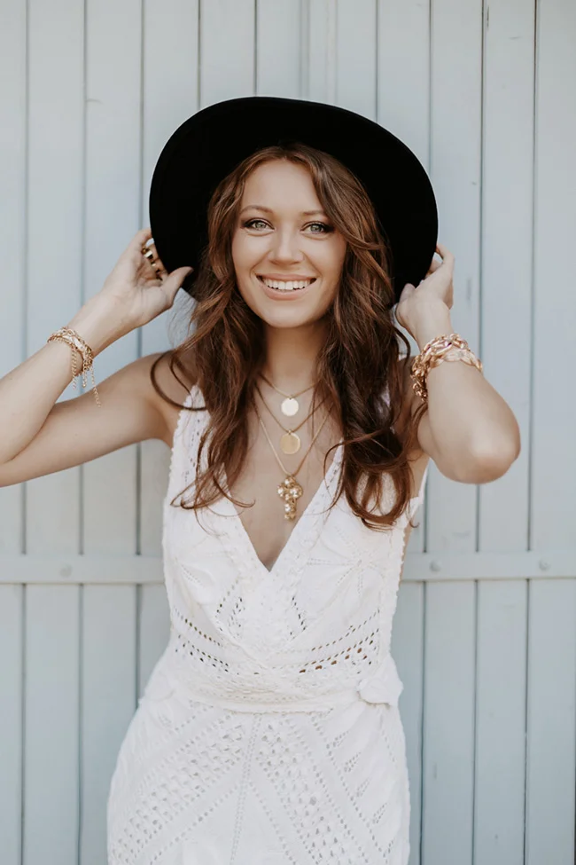Femme souriante en robe blanche dentelée et chapeau noir, look bohème chic devant un mur bleu clair.