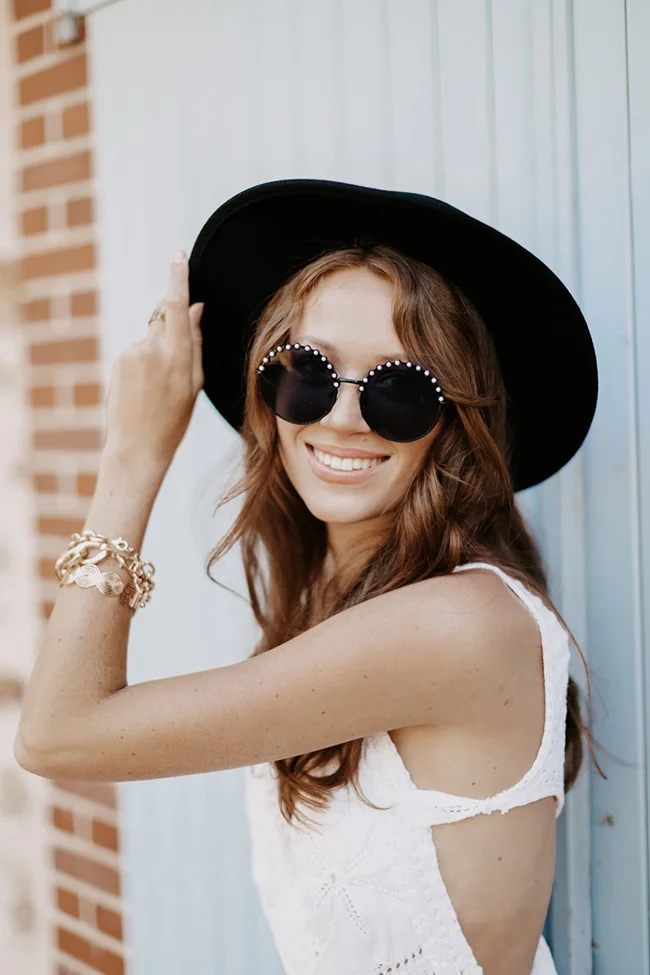 Femme souriante en robe blanche, chapeau noir et lunettes perlées, pour un look de mariée glamour et bohème.