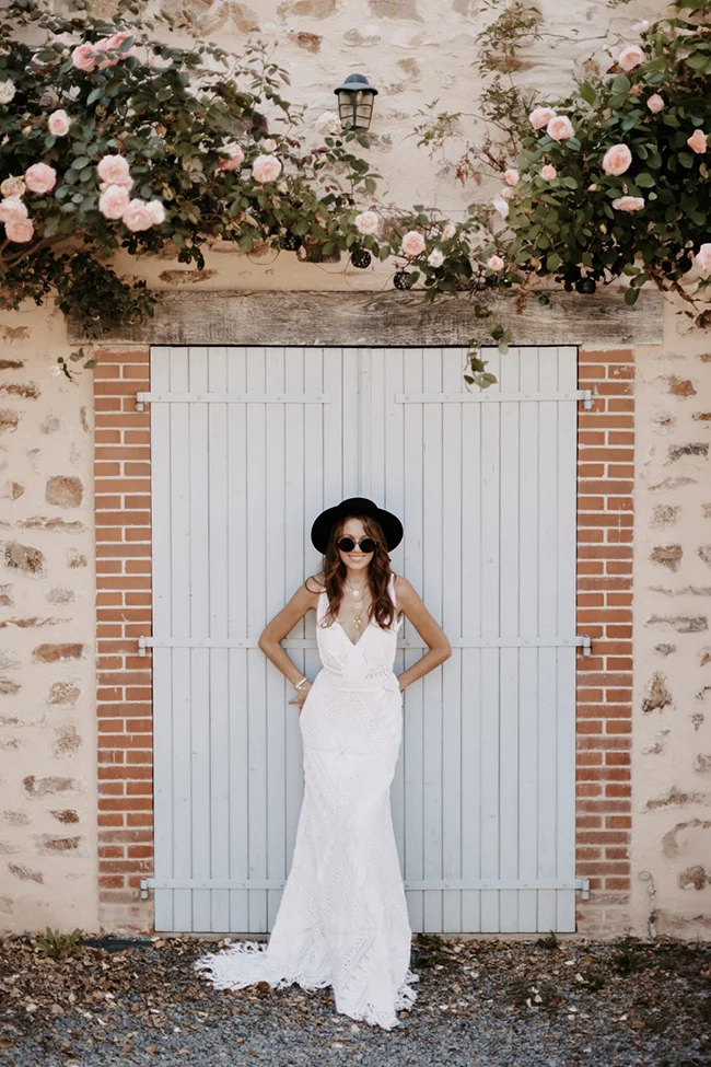 Mariée glamour en robe blanche et chapeau noir posant devant une porte rustique entourée de roses.