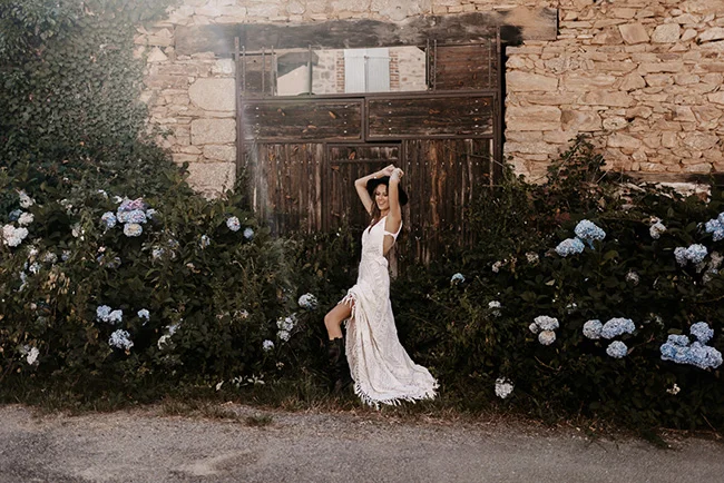 Mariée glamour en robe blanche fendue posant devant une porte rustique entourée d'hortensias bleus.