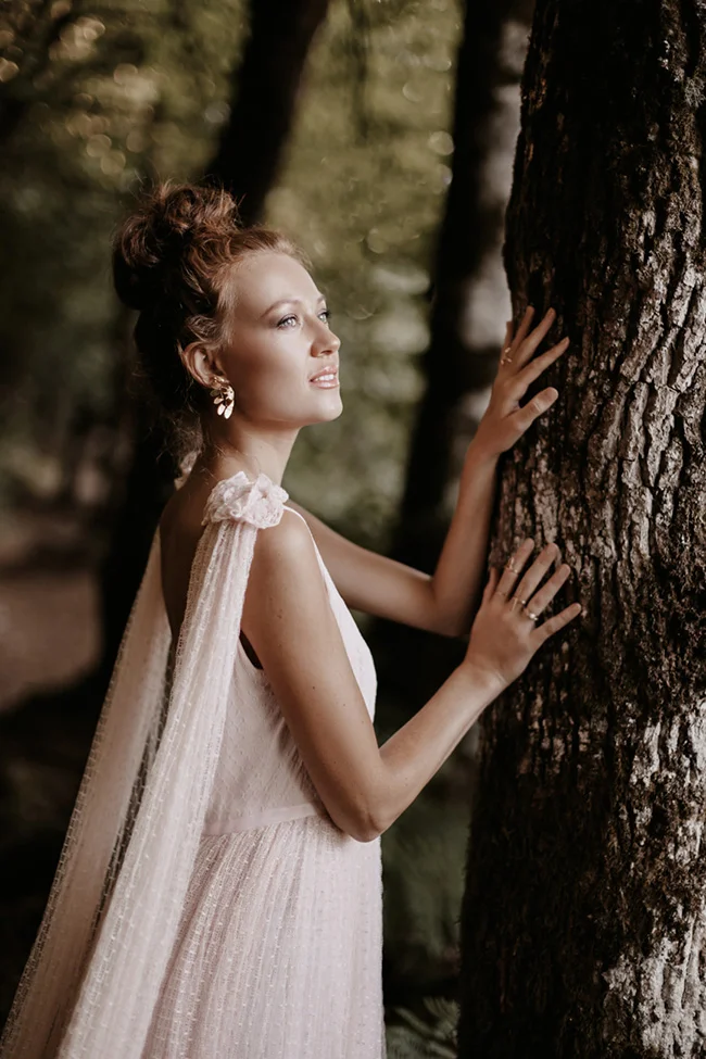 Une mariée glamour en robe rose poudré, cheveux relevés, s'appuyant avec douceur contre un tronc d'arbre en forêt.
