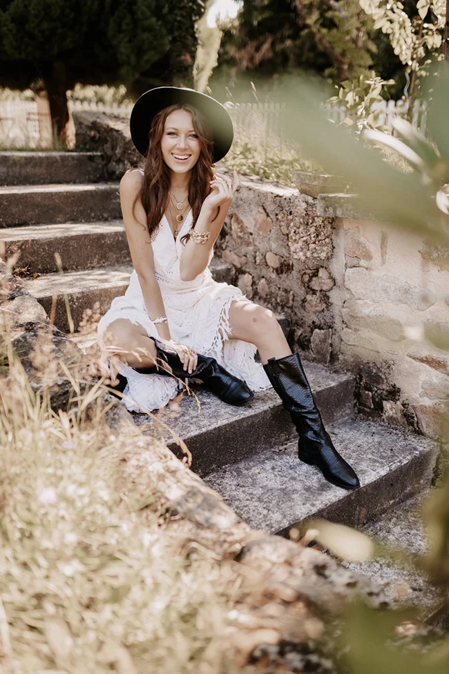 Mariée souriante en robe blanche bohème, chapeau noir et bottes en cuir, assise sur des marches en pierre ensoleillées.