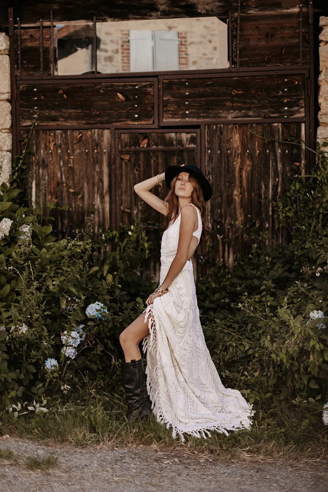 Mariée glamour en robe blanche bohème et chapeau noir, posant devant une vieille porte en bois rustique et verdoyante.