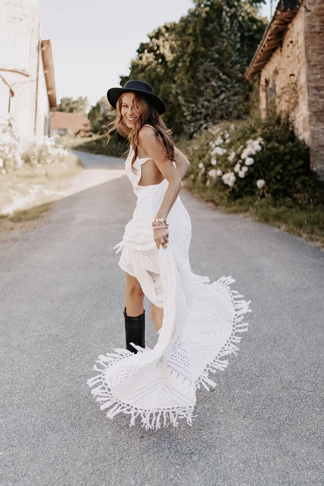 Mariée souriante en robe blanche bohème à franges, chapeau noir et bottines, posant dans une ruelle rustique.