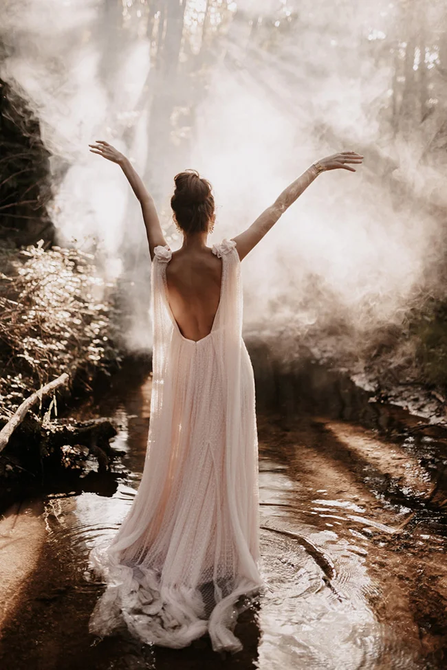 Mariée en robe fluide, bras levés dans un ruisseau brumeux et lumineux, ambiance sauvage et onirique.