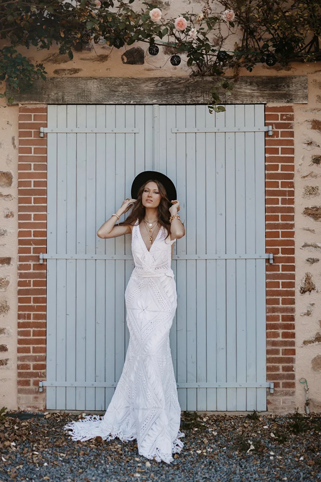 Mariée glamour en robe blanche bohème et chapeau noir, posant devant une porte bleue et un mur de briques.