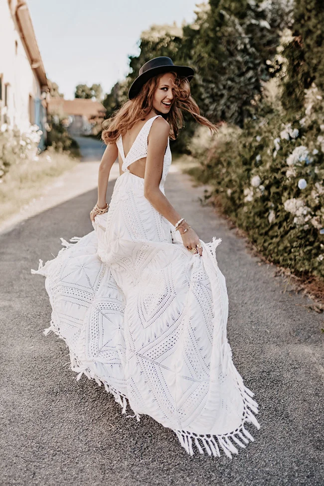 Mariée souriante en robe blanche bohème et chapeau noir, tournoyant sur un chemin champêtre.