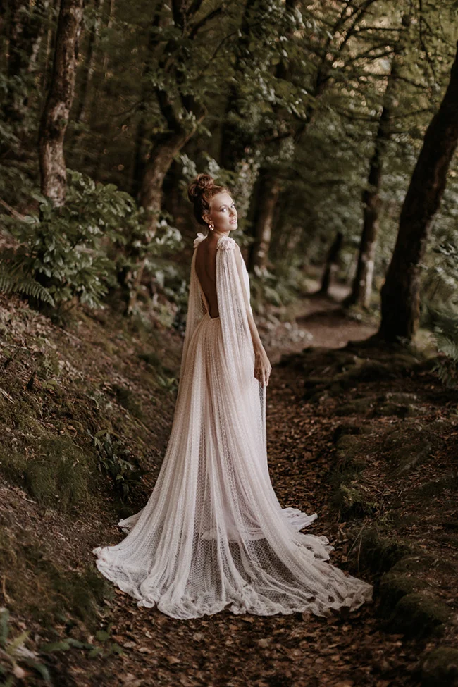 Mariée en robe blanche vaporeuse et cape en dentelle, posant avec élégance dans une forêt sombre et mystérieuse.