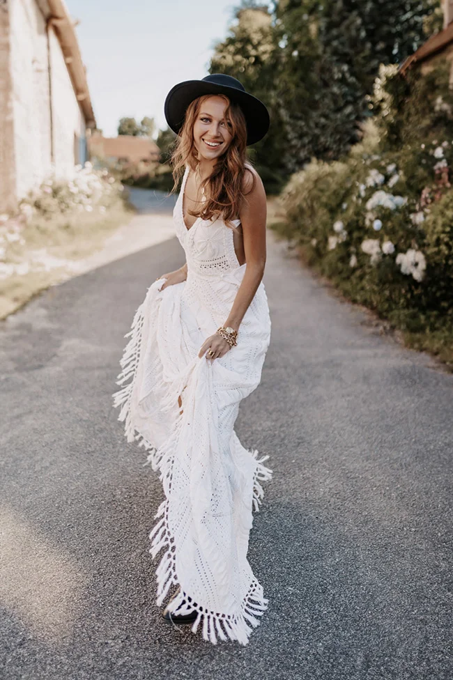 Mariée souriante en robe blanche bohème à franges et chapeau noir, posant dans une allée champêtre et lumineuse.
