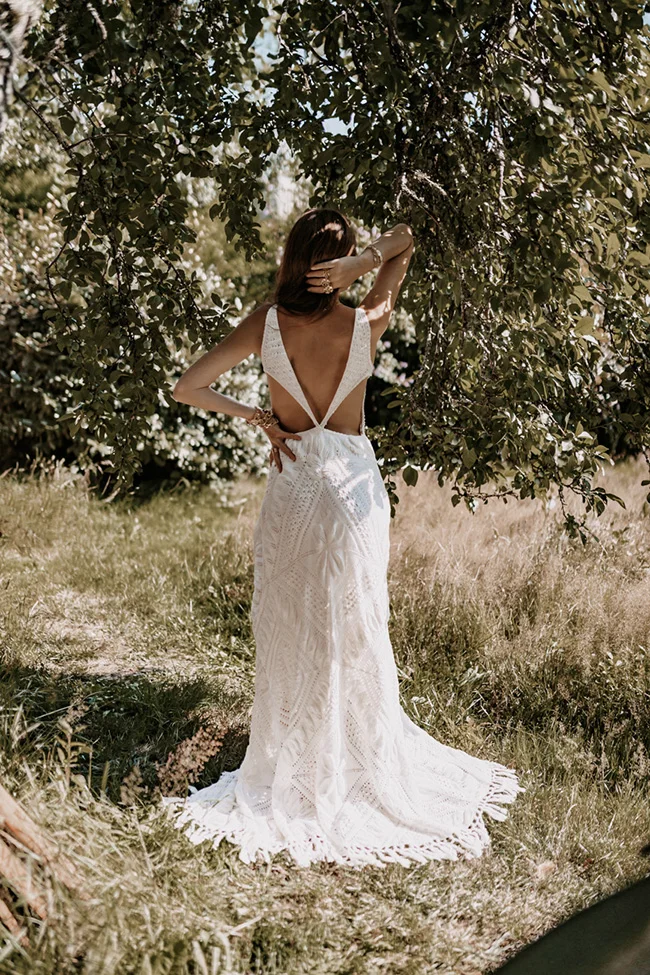 Mariée de dos en robe blanche en dentelle, posant sous un arbre dans un champ sauvage et lumineux.
